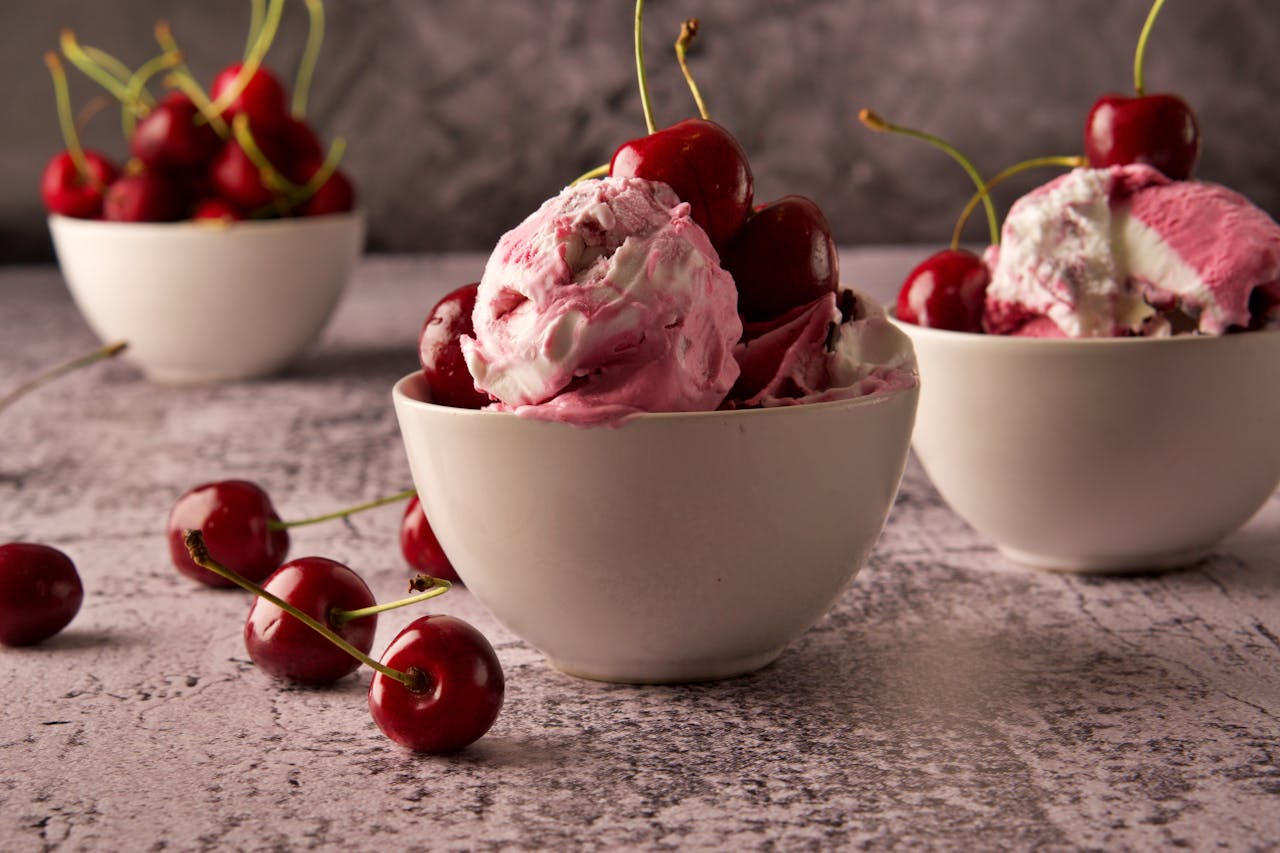 Three white ceramic bowls filled with cherry ice cream and fresh cherries on a textured surface.