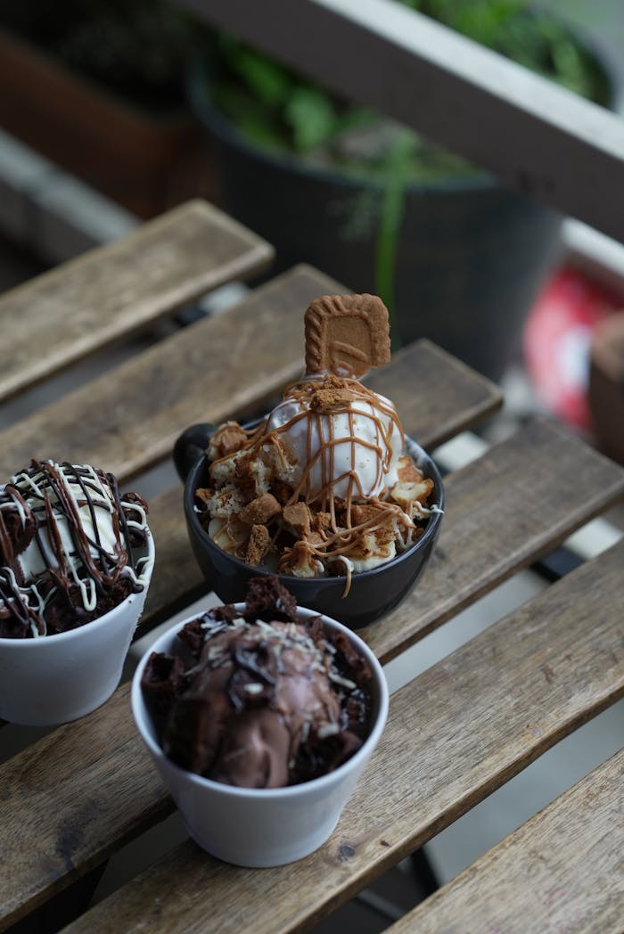 Close-up of assorted ice cream desserts with chocolate and caramel toppings on a rustic wooden table outdoors.